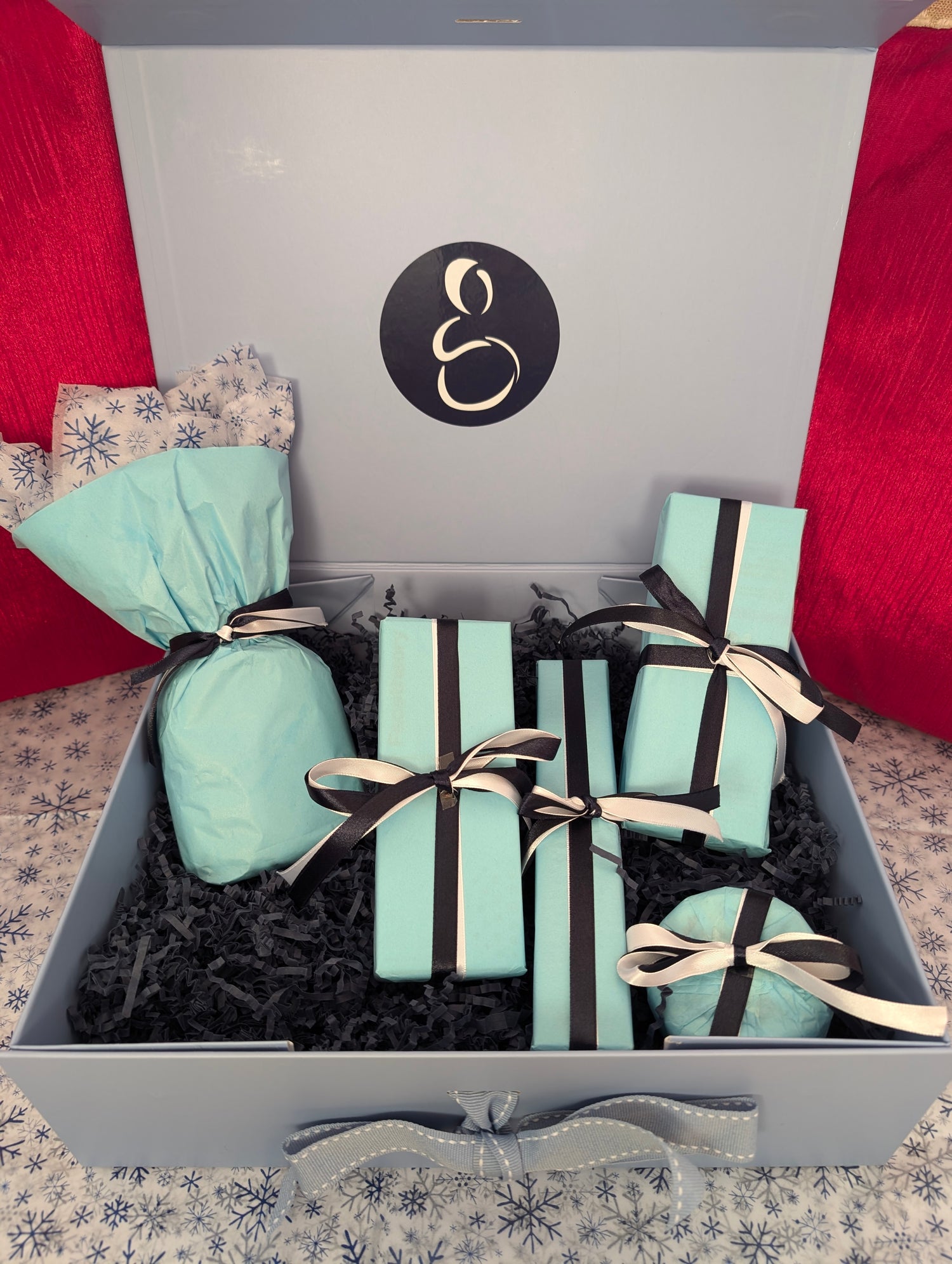 Gift box with turquoise-wrapped presents tied with black and white ribbons, placed on black crinkle paper, and displayed against a festive red and snowflake-patterned background.